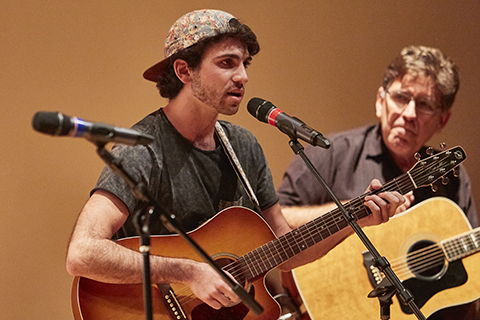 Man with a acoustic guitar performs at an event while another man in the background looks on