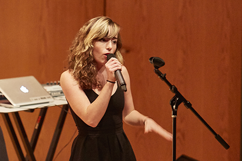 Woman with blonde hair and black dress sings into a microphone on stage